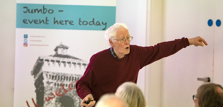 A man gestures as he talks to an audience. He is standing in front of a banner with the words event here today.