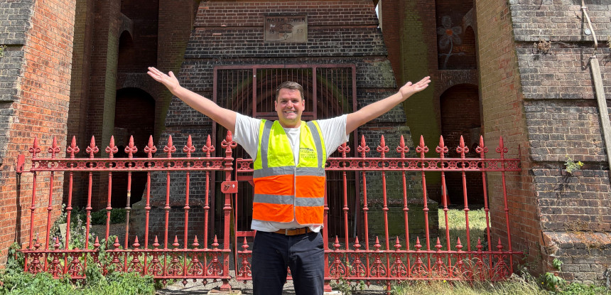 A smiling person wearing a high visibility jacket stands in front of a red brick building. Their arms are outstretched in excitement.