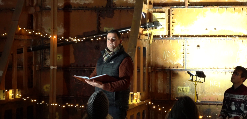 A person, wrapped in warm clothing, stands in a dimly-lit room which has rusty metal panels for walls. It is a huge water tank. The person is an actor and is reading from a book.