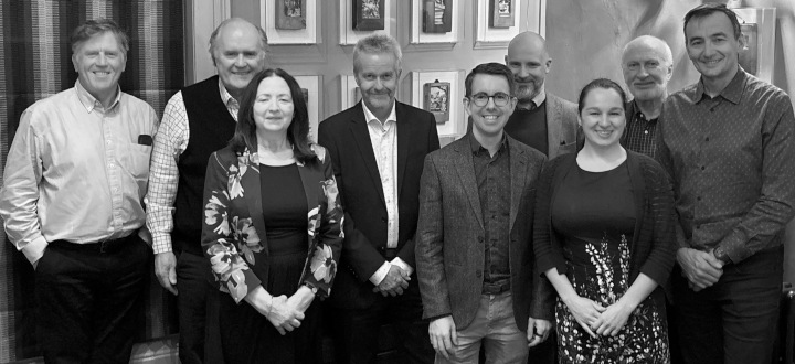 A group of nine adults, seven men and two women, pose in a relaxed manner indoors. Everyone is smiling and dressed in semi-formal attire.