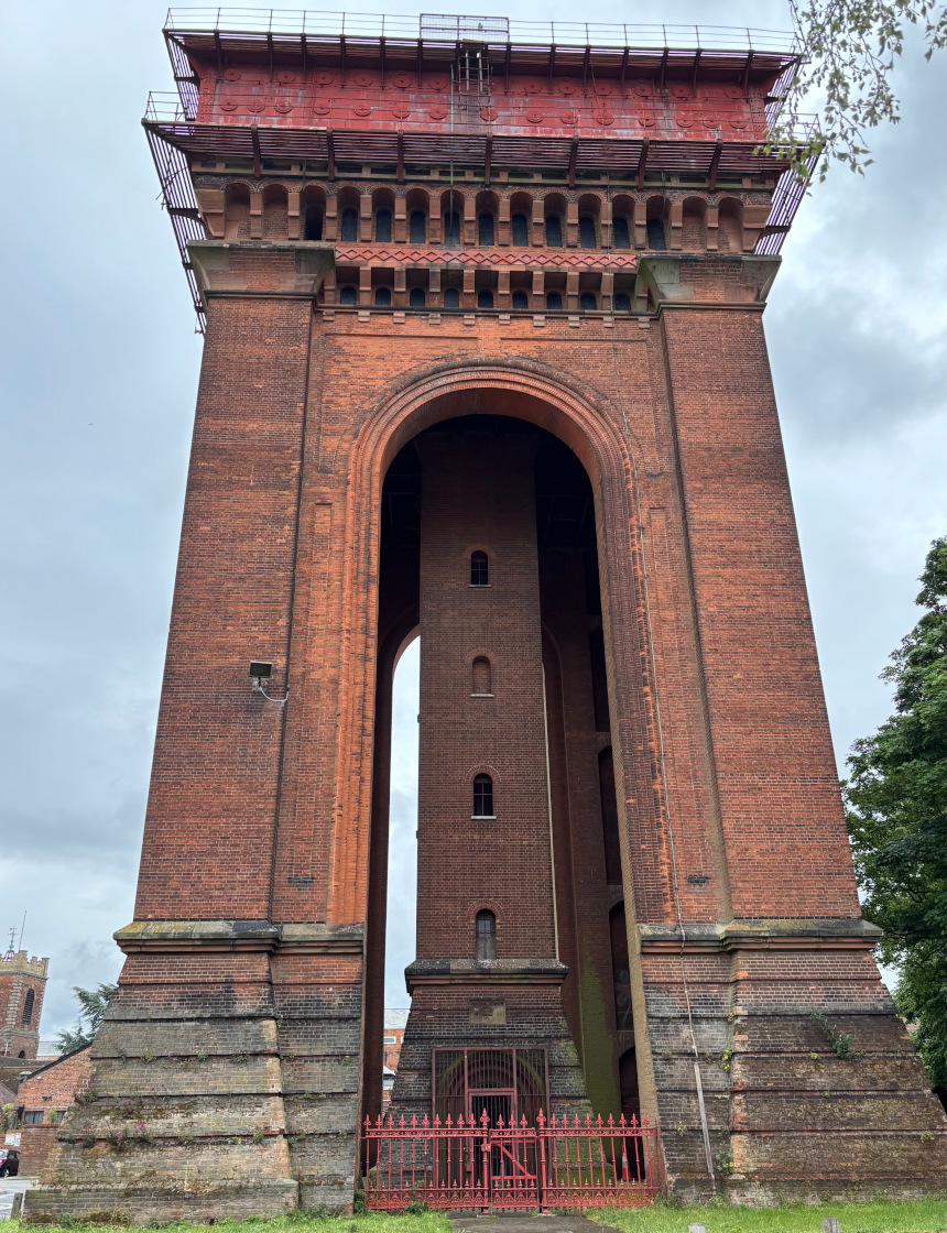 A tall building, consisting of five legs of red bricks and an arch, can be seen against a blue sky. The building is a disused water tower and is topped with a large, square red metal tank.