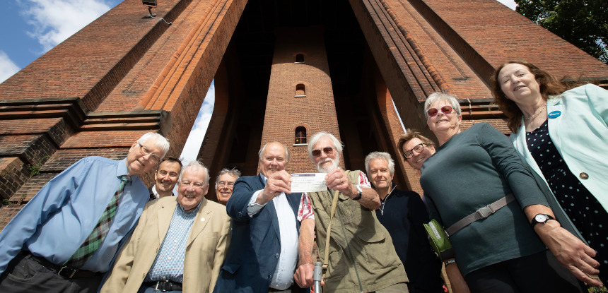 A group of smiling men and women are seen in front of a tall brick building. They are gathered around two men holding a cheque.