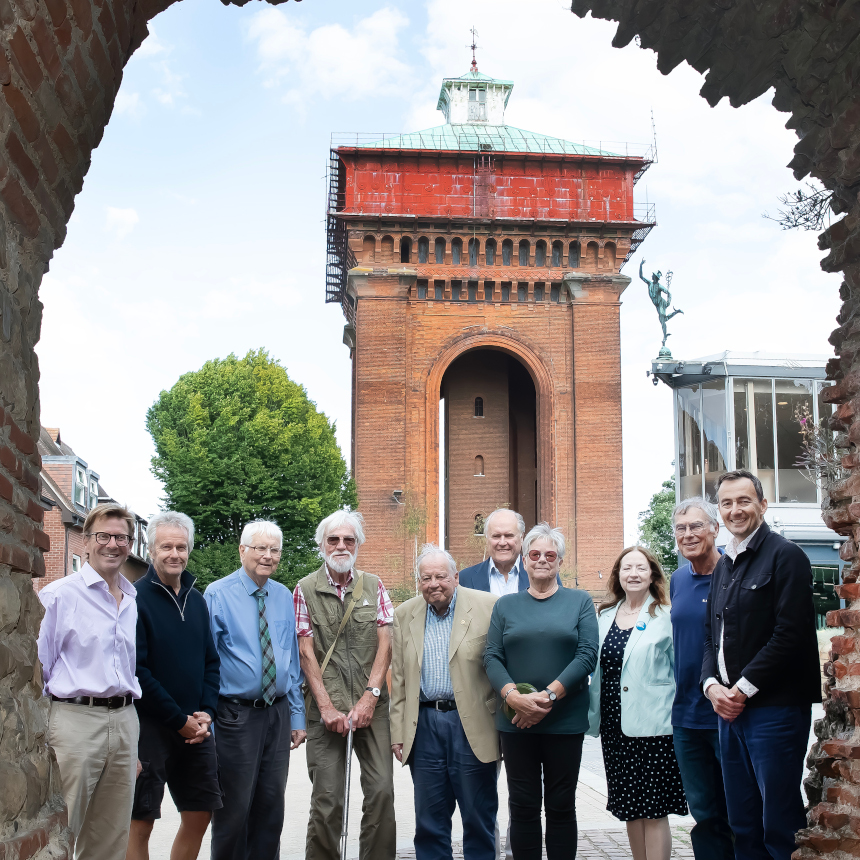 A group of smiling men and women at seen with a very tall brick building in the background. The building is a disused water tower and is topped with a large, square red metal tank.