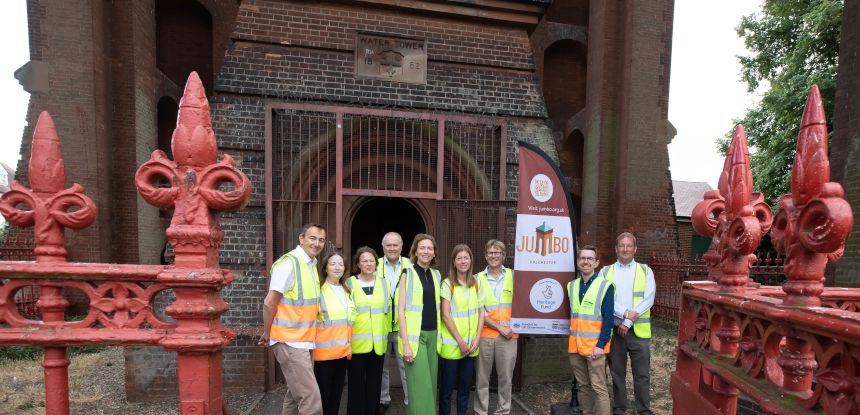 A group of smiling men and women stand in front of a brick building. The image includes ornate red iron railings.