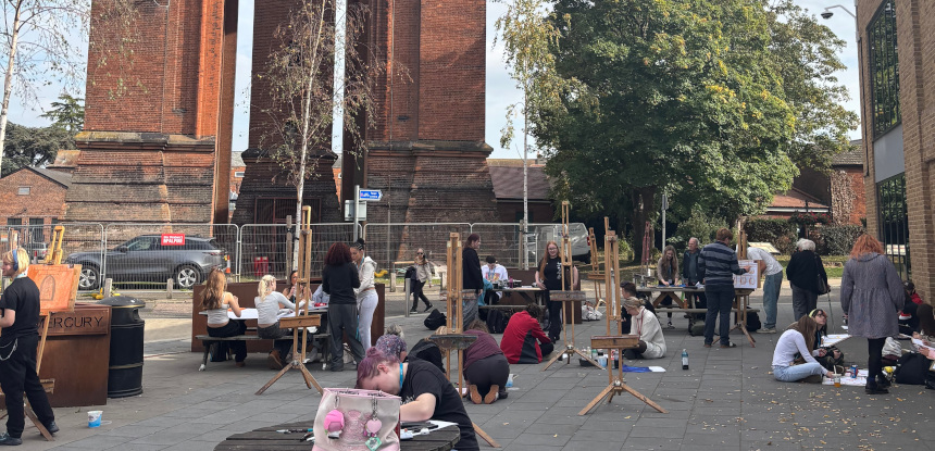 People stand at easels and sit on floor and at tables, drawing in the shadow of a red brick building. People stand at easels and sit on floor and at tables, drawing in the shadow of a red brick building.