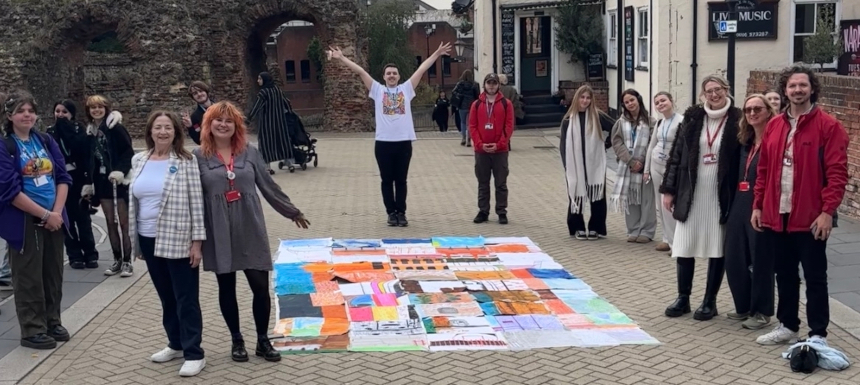 A group of people stand around a large and colourful drawing on a pavement. The drawing is made up of individual sheets of paper. A group of people stand around a large and colourful drawing on a pavement. The drawing is made up of individual sheets of paper.