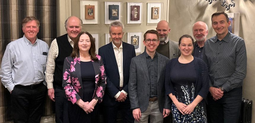 A group of nine adults, seven men and two women, pose in a relaxed manner indoors. Everyone is smiling and dressed in semi-formal attire.