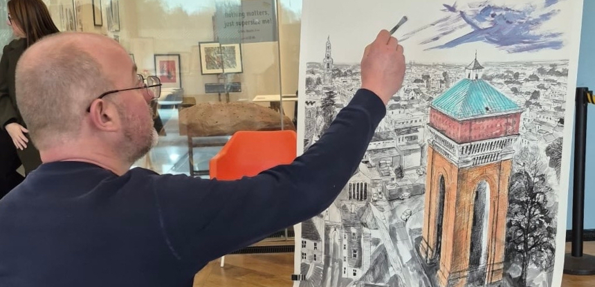 A man in glasses sketches a detailed cityscape, focusing on a tall, red-brick tower with a teal roof. The setting is a modern room with art on the walls.