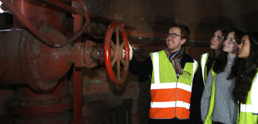 Three young people are being shown huge red coloured iron pipes and values by a smiling man in a high viz vest.