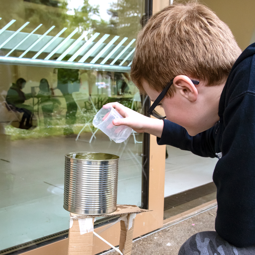 A young person pours water from a plastic cup into a tin can balanced on top of a cardboard tower.