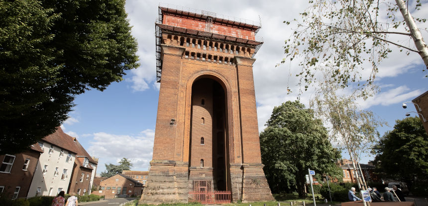 A tall red brick building with a red metal water tank on top is pictured on a sunny day.