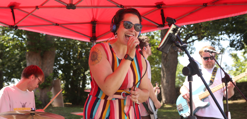 A person in a bright dress sings as they stand under a large red gazebo. Behind them is a drummer and a guitarist.