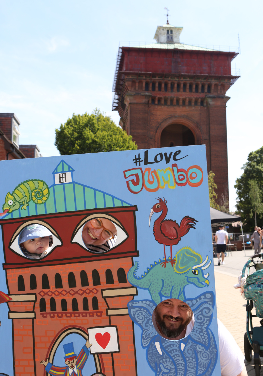 A huge red brick tower with a red water tank on top can be seen. In the foreground, two smiling adults and a small child peep through holes in a large, colourful artwork depicting the tower and exotic animals.