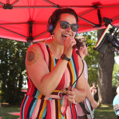 A person in a bright dress sings as they stand under a large red gazebo. Behind them is a drummer and a guitarist. 