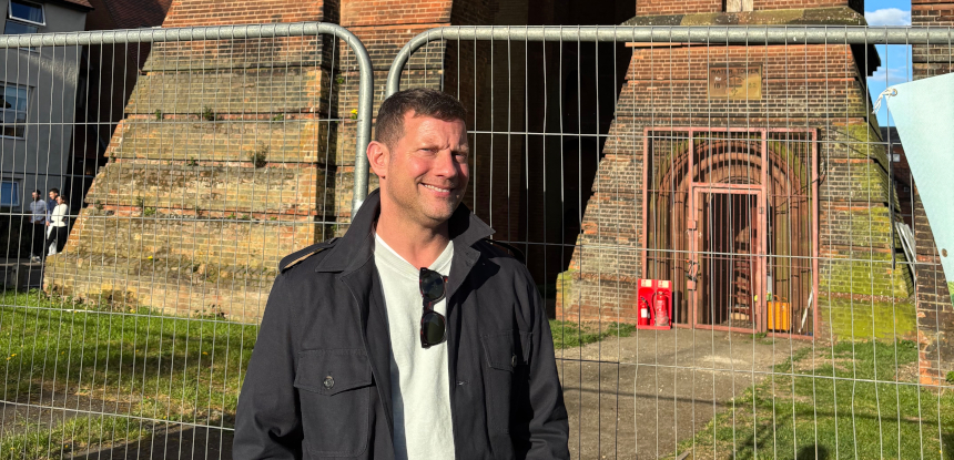 Dermot O'Leary stands smiling in front of the path to a red brick tower