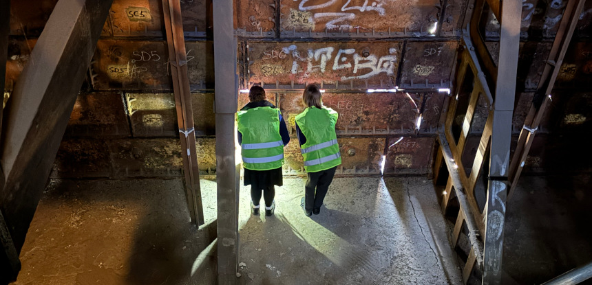 Two people wearing high visibility jackets look through the gaps of a metal tank next to a diagonal support.