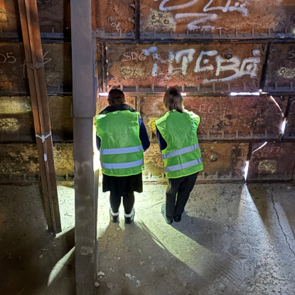 Two people wearing high visibility jackets look through the gaps of a metal tank next to a diagonal support.